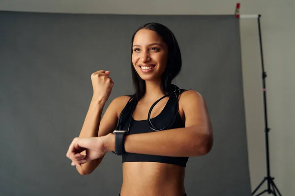 Happy young biracial woman wearing sports bra with raised fist holding up fitness tracker in studio