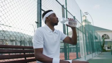 Young black man in sportswear taking a break and drinking water during tennis practice at sports court