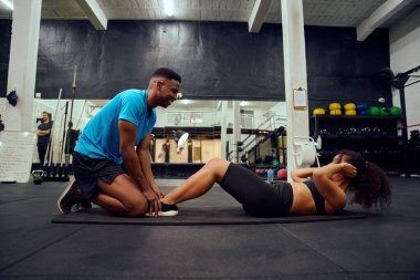 Multi-ethnic friends doing cross fit in the gym. African American male encouraging African American female during sit-ups. High quality photo