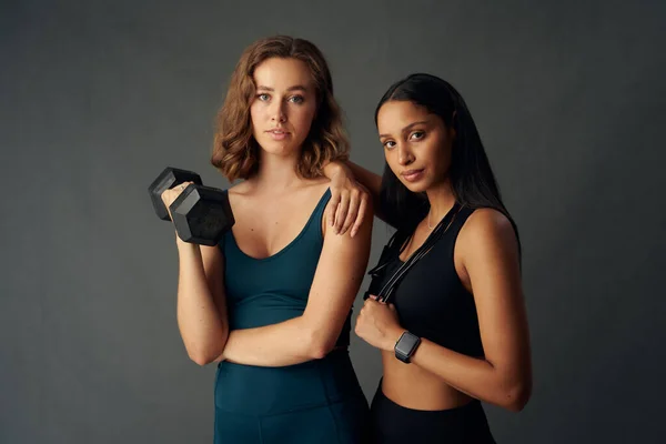 Determined young woman with friend wearing sports clothing looking at camera while holding dumbbell