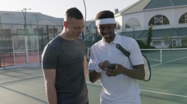 Happy young men in sportswear talking while using mobile phone during tennis practice at sports court