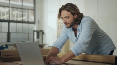 Ofiste dizüstü bilgisayarda yazarken ürünün üzerine eğilen beyaz tişört giyen odaklanmış genç bir adam.