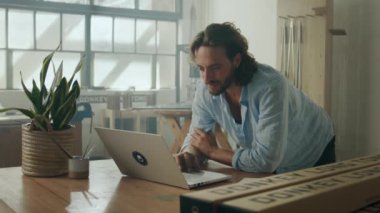 Ofisteki ürünlerin yanında dizüstü bilgisayarda yazarken dirseklerine yaslanan beyaz gömlekli genç bir adam.