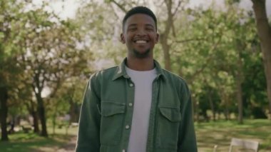 Tişört giyen mutlu genç adam parkta kolları bağlı kameraya bakıyor.