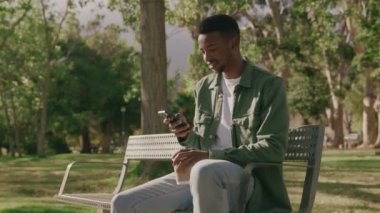 Parkta bankta otururken cep telefonu kullanan ve kahve içen genç siyah bir adam.