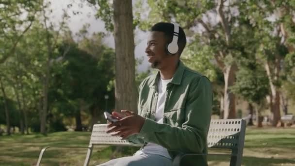 Genç Siyahi Adam Gülerek Kulaklıkla Ağaç Altındaki Bankta Müzik Dinleyerek — Stok video