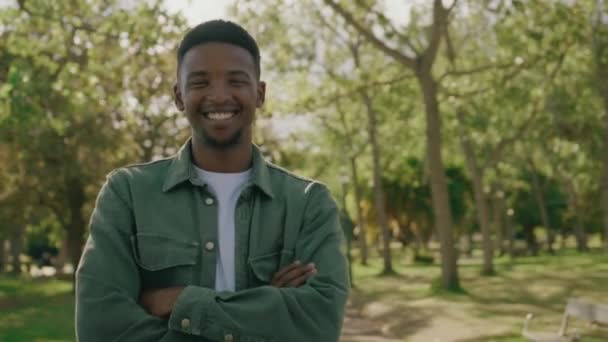 公園の木の下で腕を組んでカメラを見て笑ってシャツを着ている若い黒人男性 — ストック動画