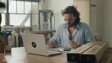 Gömlekli genç beyaz adam dizüstü bilgisayar kullanıyor ve ofisteki ürünlerin yanında cep telefonu kullanıyor.