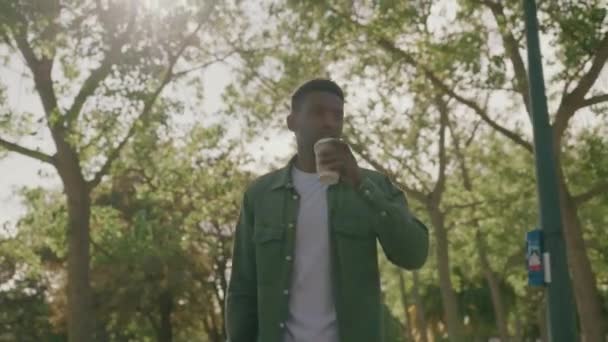 公園の木の下の使い捨てカップにシャツを着て歩くとコーヒーを飲む若い黒人男性 — ストック動画