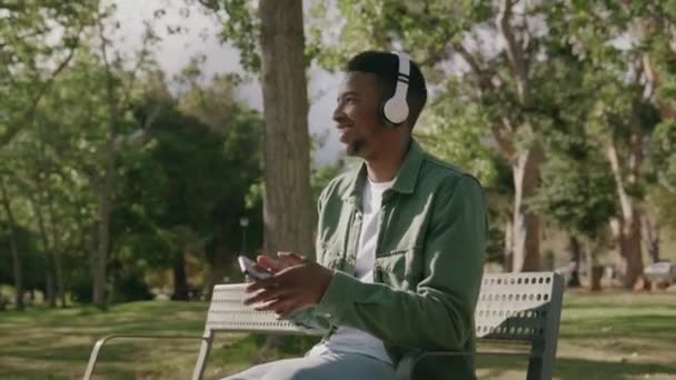 身穿休闲装的黑人年轻人笑着坐在树下的长椅上 用耳机听音乐 — 图库视频影像