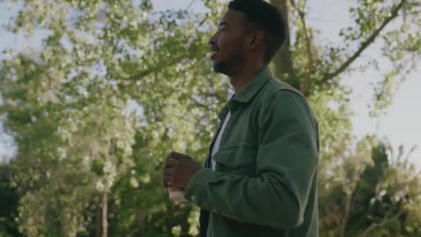 公園の木の下で使い捨てカップでコーヒーと一緒に歩いてシャツを着て幸せな若い黒人男性 — ストック動画