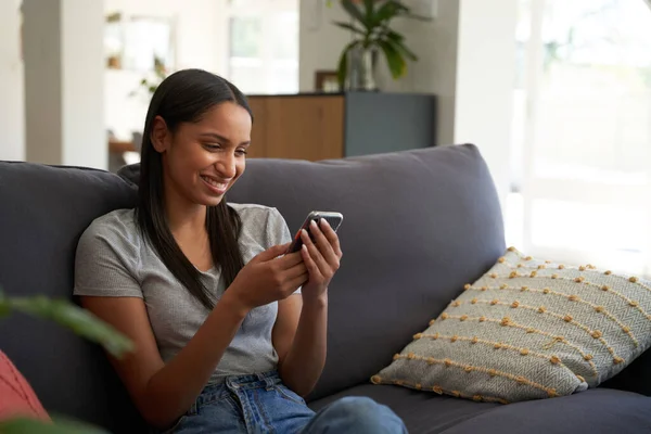 在家里沙发上使用手机时穿着休闲装 面带微笑的年轻白人妇女 — 图库照片