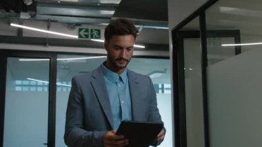 Şirketin koridorunda yürürken dijital tablet kullanan, iş hayatında odaklanmış beyaz bir adam.