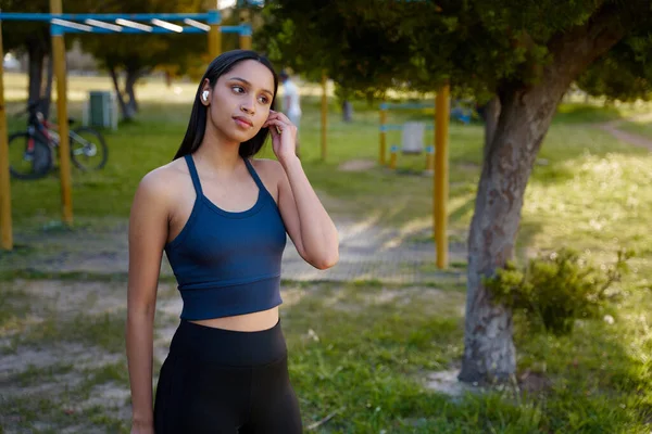 公園のワイヤレスイヤホンで音楽を聴くスポーツウェアを着ている若い女性 — ストック写真