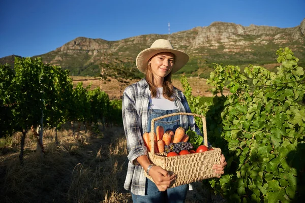 Contented young caucasian woman wearing casual clothing carrying basket with fresh harvest on farm