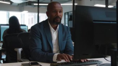 Tişörtlü ve spor ceketli yetişkin bir adam şirket ofisindeki masada bilgisayarda daktilo kullanıyor.