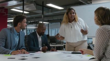 İş kıyafetlerindeki çok ırklı iş arkadaşları ofiste toplantı sırasında beyaz tahtayla konuşup el kol hareketi yapıyorlar.