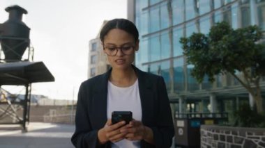 İş kıyafetleri giyen çok ırklı genç bir kadın caddedeki ofis binasının yanındaki cep telefonunda daktilo kullanıyor.
