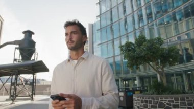İş kıyafetleri giyen genç beyaz adam caddedeki ofis binasının yanındaki cep telefonunda daktilo kullanıyor.