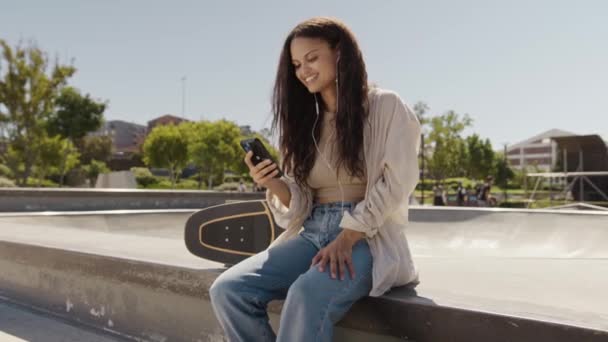 スケートパークで携帯電話を使用してカジュアルな服を着ている幸せな若い異人種の女性 — ストック動画