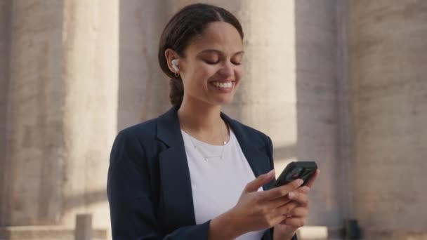 建物の隣に携帯電話を使用してビジネスウェアを身に着けている幸せな若い異人種の女性 — ストック動画