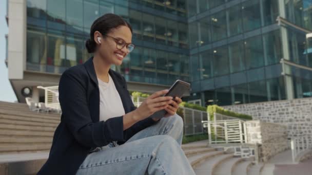 ストリートのオフィスの隣にデジタルタブレットでステップに座っているビジネスウェアの幸せな若い異人種の女性 — ストック動画