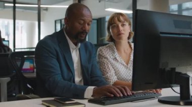 Yetişkin bir adam ve genç bir kadın bilgisayarda yazı yazıyor ve şirket ofisindeki masada konuşuyorlar.