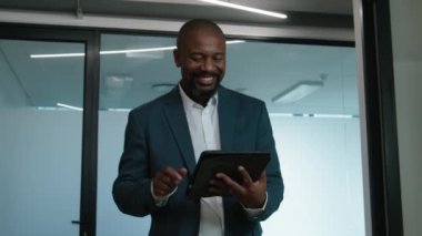 Şirketin koridorunda yürürken dijital tablet kullanan mutlu, olgun siyahi bir adam.