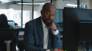 İş hayatında olgun siyah bir adam şirket ofisindeki masada bilgisayar başında yazarken kaşlarını çatıyor.