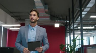 Elinde dijital tablet tutan genç adam şirket ofisindeki iş arkadaşlarına el sallıyor.