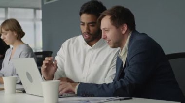 İş kıyafetleri giyen dört genç iş adamı ofiste dizüstü bilgisayarla konuşuyor ve el kol hareketi yapıyor.
