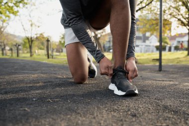 Spor giysileri giyen genç siyahi adamın parkta ayakkabı bağcığı bağlarken diz çöktüğü yakın plan.