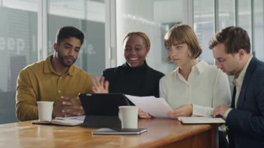 İş kıyafetleri giyen dört yetişkin şirket ofisinde toplantı sırasında dijital tablet kullanarak video görüşmesi yapıyor.