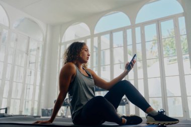 Spor kıyafetleri giyen, spor salonunda oturan ve cep telefonu kullanan çok ırklı genç bir kadının profili.