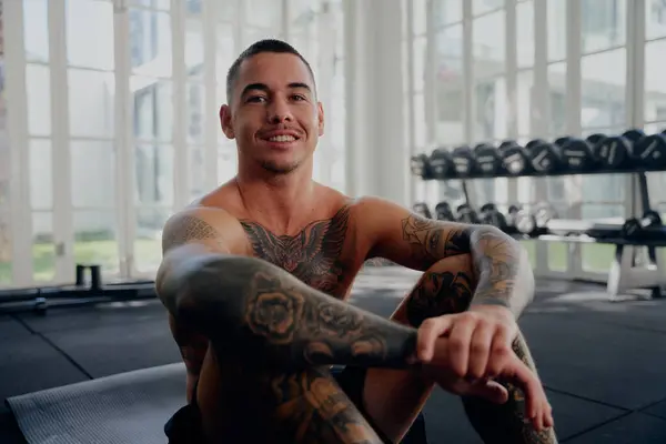 Happy shirtless young tattooed multiracial man wearing shorts sitting while taking a break at the gym
