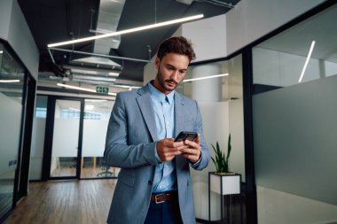 İş kıyafetleri giyen genç beyaz adam şirket binasının koridorunda cep telefonu kullanıyor.