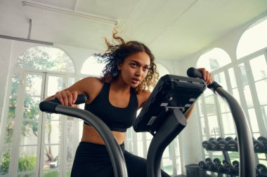 Spor kıyafetleri giyen, spor salonunda bisiklete binen odaklanmış, çok kültürlü bir kadın.
