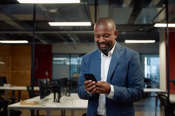 Happy mature black man in businesswear using mobile phone in corporate office