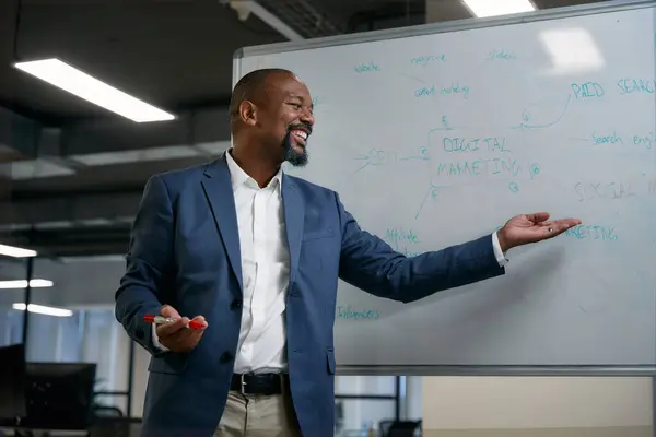 ハッピー成熟した黒人ビジネスマン笑顔とコーポレートオフィスでのミーティング中にホワイトボードの隣にジェスチャー — ストック写真