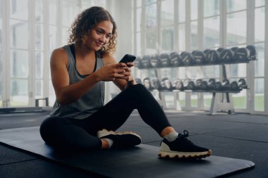 Spor kıyafetleri giyen, spor salonunda dinlenirken cep telefonu kullanan çok ırklı genç bir kadın.