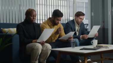 Genç çok ırklı yetişkinler iş kıyafetlerini giyip kanepede oturup şirket ofisinde evrak okuyorlar.