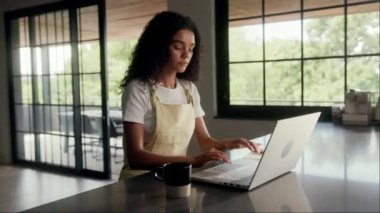 Bir kadın, bir binanın içindeki pencerenin yanındaki ahşap masada dizüstü bilgisayar kullanarak oturuyor. İç tasarımda sert ahşap döşeme ve cam elementler yer alıyor.
