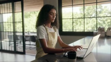 Bir kadın, bir evin penceresinin yanındaki ahşap bir masada dizüstü bilgisayar kullanarak oturuyor. Kalçası ahşap zeminde duruyor.