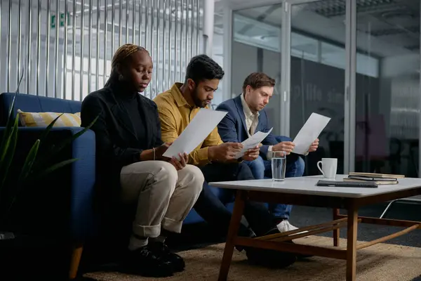 ソファーに座り オフィスで文書を読んでいるビジネスウェアを身に着けている3人の多種多様な若者 ロイヤリティフリーのストック写真