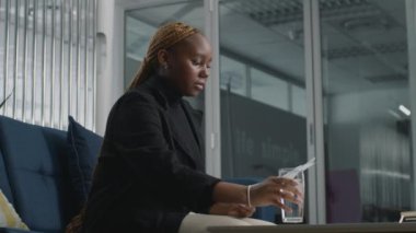 İş kıyafetleri giyen, su içen ve iş arkadaşını beklerken kitap okuyan genç bir kadın.