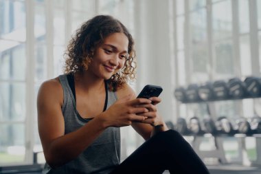 Spor kıyafetleri giyen, spor salonunda mola verirken cep telefonuyla mesajlaşan çok ırklı genç bir kadın.