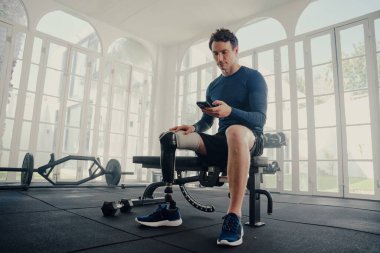 Yapay bacaklı, genç, beyaz bir adam spor salonunda cep telefonuyla göğüs presi yapıyor.