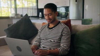Sakallı bir adam kanepede oturuyor ve dizüstü bilgisayar kullanıyor. Bir pencere ev etkinliğini gösterirken, başparmağı klavyede yazı yazıyor. Eğleniyor gibi görünüyor.