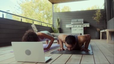 Bir erkek ve bir kadın balkonda ahşap döşemeli yoga yapıyorlar, dizüstü bilgisayara bakıyorlar. Arkaplandaki ağaç ve bina etkinliğin boş ve eğlenceli kısmına eklenir