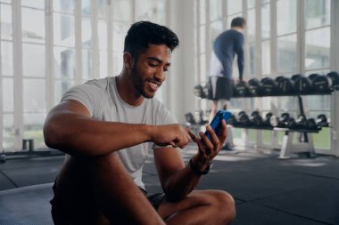 Spor kıyafetleri giyen, spor salonundaki ağırlıkların yanında cep telefonu kullanan iki mutlu çok ırklı genç adam.